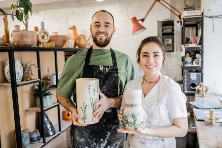 Smiling couple of artisans in aprons holding clay vases and looking at camera in ceramic workshop