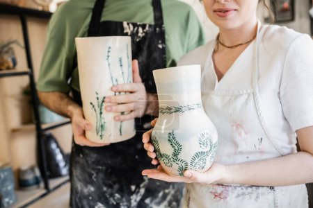 Téléchargez les photos : Vue recadrée de l'artisan dans le tablier tenant vase d'argile près de copain flou dans l'atelier de céramique - en image libre de droit
