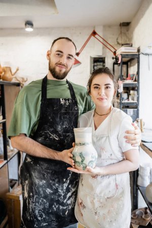 Foto de Pareja sonriente de artesanos en delantales abrazando y sosteniendo jarrón de arcilla en taller de cerámica en el fondo - Imagen libre de derechos