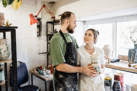 Positives Bildhauerpaar in Schürzen hält Tonvase in der Hand und spricht in Keramikwerkstatt