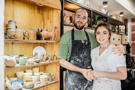 Smiling bearded craftsman in apron hugging girlfriend and looking at camera in ceramic workshop