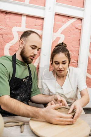 Foto de Artesano barbudo en delantal formando tazón de barro y hablando con su novia en taller de cerámica - Imagen libre de derechos