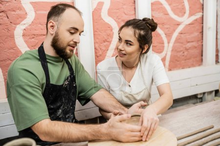 Téléchargez les photos : Artisanat souriant dans tablier parler à petit ami et faire bol d'argile ensemble dans l'atelier de céramique - en image libre de droit