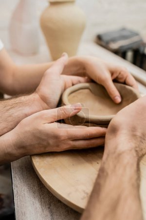 Cropped view of couple of artisan shaping clay bowl near table in ceramic workshop
