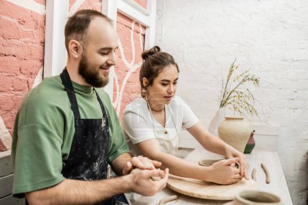 Artesana en delantal talkig a novio y moldeando tazón de barro en taller de cerámica