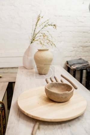 Clay bowl on wooden board near vases with flowers on table in ceramic workshop