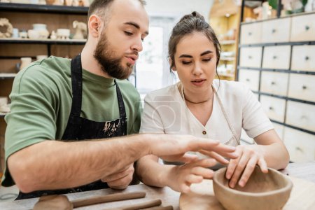 Artesano barbudo en delantal formando tazón de barro y hablando con su novia en taller de cerámica
