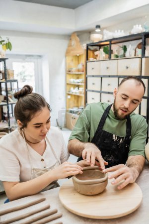 Téléchargez les photos : Couple de potiers dans des tabliers façonnant bol d'argile ensemble tout en travaillant dans un atelier de céramique - en image libre de droit