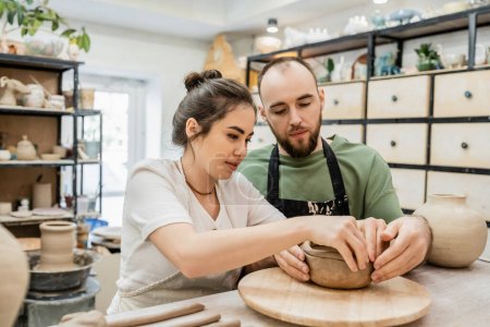Téléchargez les photos : Brunette artisan dans tablier faisant bol d'argile avec petit ami ensemble dans l'atelier de céramique - en image libre de droit