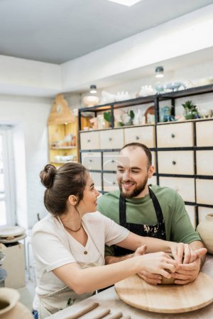 Téléchargez les photos : Couple souriant d'artisans dans des tabliers faisant bol d'argile et se regardant dans un studio de céramique - en image libre de droit