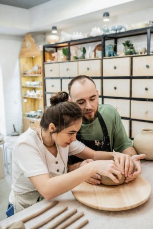 Brunette craftswoman in apron crafting clay bowl with boyfriend on wooden board in studio