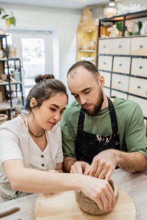 Téléchargez les photos : Brunette artisan en tablier façonnant bol d'argile avec petit ami en atelier de céramique à l'arrière-plan - en image libre de droit