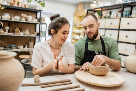 Foto de Artesana sonriente en delantal moldeado de arcilla mientras novio haciendo tazón en taller de cerámica - Imagen libre de derechos