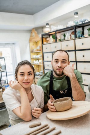 Lächelnde Kunsthandwerkerin in Schürze blickt in die Kamera neben traurigem Freund und Ton in Keramikwerkstatt