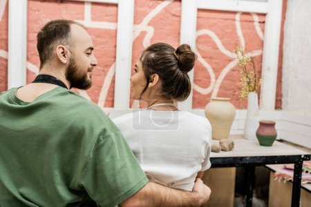 Foto de Artesano barbudo en delantal abrazando novia mientras está sentado cerca de la arcilla en el taller de cerámica - Imagen libre de derechos