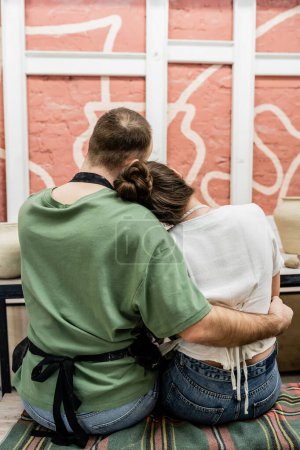 Rückansicht von Handwerker in Schürze, der Freundin umarmt, während er neben Ton im Töpferatelier sitzt