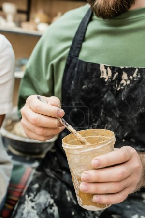 Cropped view of craftsman in apron holding clastic cup with dye and paintbrush in ceramic workshop