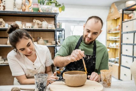 Sourire artisan colorant bol d'argile près de petite amie dans l'atelier de céramique à l'arrière-plan