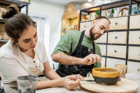 Bärtiger Handwerker in Schürze färbt Keramikschale mit verschwommener Freundin in Keramikwerkstatt