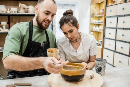 Téléchargez les photos : Artisanat souriant dans un bol en argile à colorier avec fiancée dans un atelier en céramique à l'arrière-plan - en image libre de droit