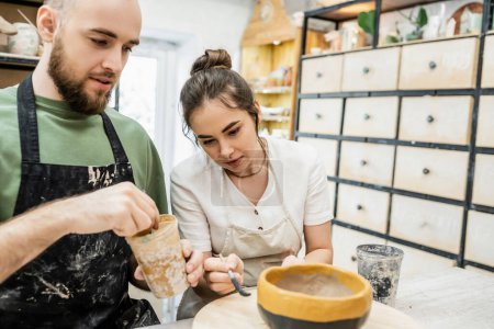 Téléchargez les photos : Artisanat dans un tablier colorant bol en argile avec petite amie floue ensemble dans un atelier de céramique - en image libre de droit