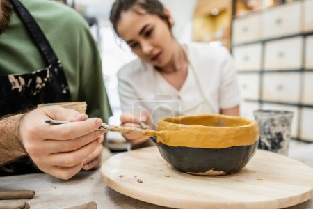 Cropped view of couple of artisans coloring ceramic bowl together in pottery studio, banner