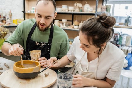 Bearded craftsman in apron dying clay bowl with girlfriend and talking in ceramic workshop
