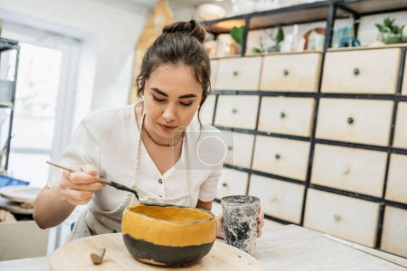 Artisanat Brunette dans un tablier colorant bol en céramique tout en travaillant dans un atelier de poterie en arrière-plan