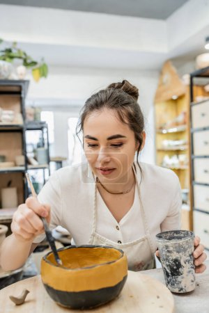Téléchargez les photos : Brunette artisan tenant tasse en plastique tout en mourant bol d'argile tout en travaillant dans un atelier de poterie - en image libre de droit