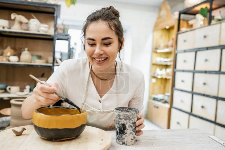 Téléchargez les photos : Joyeux artisan brune coloration de l'argile céramique tout en travaillant dans l'atelier de poterie en arrière-plan - en image libre de droit