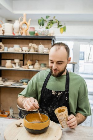 Joyful male potter in apron coloring unglazed clay bowl on wooden board in ceramic studio