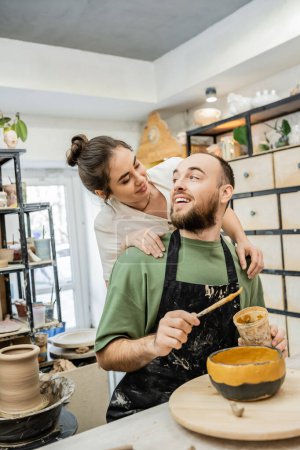 Photo for Positive craftswoman in apron hugging and looking at boyfriend coloring clay bowl in pottery studio - Royalty Free Image