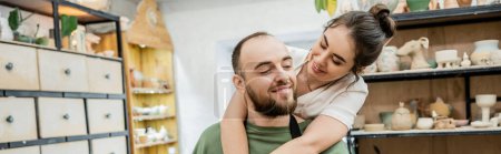 Foto de Artesana sonriente en delantal abrazando novio barbudo y trabajando en taller de cerámica, pancarta - Imagen libre de derechos