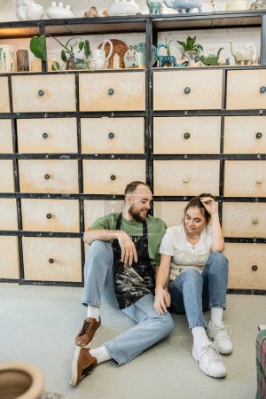Lächelndes Kunsthandwerkerpaar in Schürzen im Gespräch, während es in der Keramikwerkstatt neben Schrank sitzt