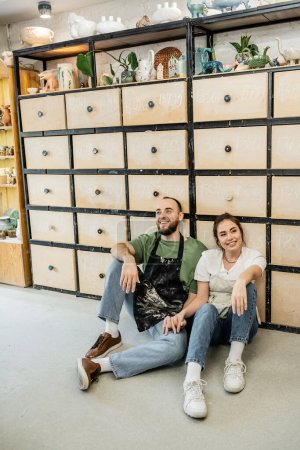 Téléchargez les photos : Couple agréable d'artisans dans des tabliers assis près de placard en atelier de céramique à l'arrière-plan - en image libre de droit