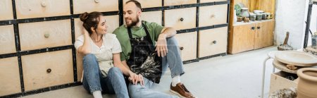 Téléchargez les photos : Couple souriant de sculpteurs assis sur le sol près du placard dans un atelier de céramique, bannière - en image libre de droit