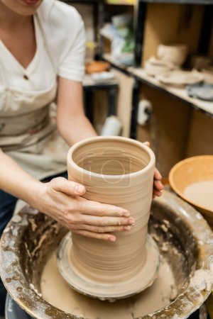 Vista recortada de alfarero hembra esculpir arcilla en la rueda de cerámica en taller de cerámica borrosa