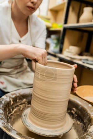 Vista recortada de la artesana en delantal moldeando arcilla sobre rueda de cerámica en taller borroso