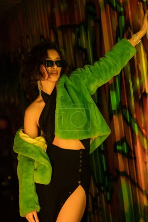 Fashionable woman in sunglasses, dress and faux fur jacket standing near graffiti in night club