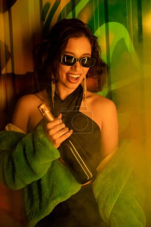 Brunette woman in sunglasses and fake fur jacket holding champagne near graffiti in night club