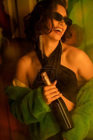 Joyful woman in sunglasses and faux fur jacket holding champagne bottle near graffiti in night club