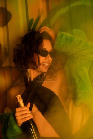 Stylish cheerful woman in sunglasses holding champagne near graffiti and smoke in night club