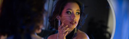 Stylowa młoda azjatycka kobieta z makijażem patrząc w pobliżu lustra i neon światło w klubie nocnym, sztandar