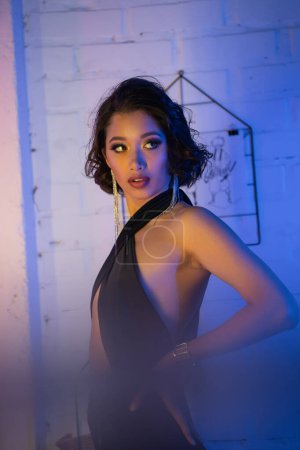 Femme asiatique à la mode avec maquillage tenant la main sur la hanche et debout dans une boîte de nuit avec lumière au néon