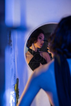 Stilvolle Asiatin im Kleid macht Selfie auf Smartphone in der Nähe von Spiegel in Nachtclub mit Neonlicht