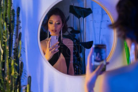 Verschwommene junge Asiatin in stylischem Outfit macht Selfie auf Smartphone neben Spiegel in Nachtclub