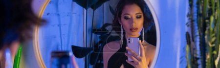 Trendy young asian woman taking selfie on smartphone near mirror and plants in night club, banner