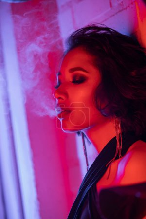 Fashionable young asian woman with makeup exhaling smoke while standing in neon light in night club