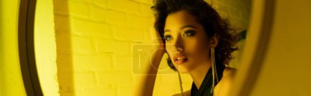 Stylish young asian woman with makeup looking at mirror in neon light in night club, banner