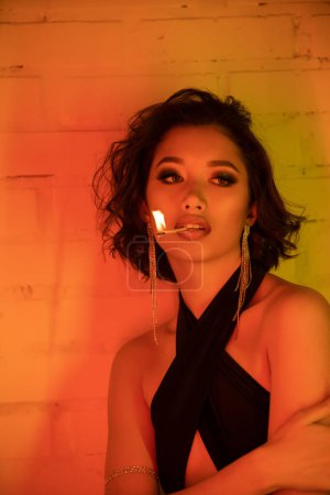 Photo for Trendy young asian woman in dress holding match with fire in lips in night club with neon light - Royalty Free Image